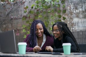Two smiling young women working with various electronic devices outdoors.